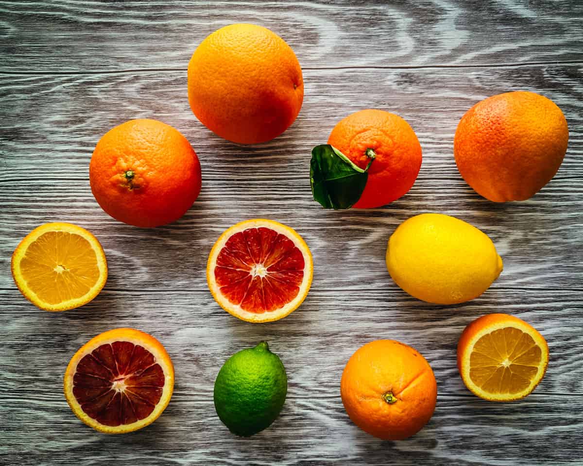 Oranges, lemons, limes, and blood oranges, some whole and some cut in half on a gray wood surface. 