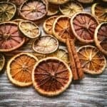 Dried slices of various citrus lying on a gray wood surface, with cinnamon sticks on top.