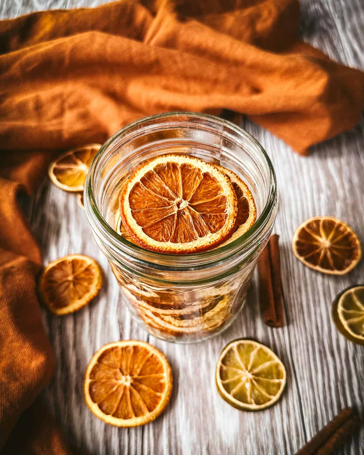 Dried citrus slices in a jar on a light wood surface, surrounded by dried citrus slices and an orange cloth.