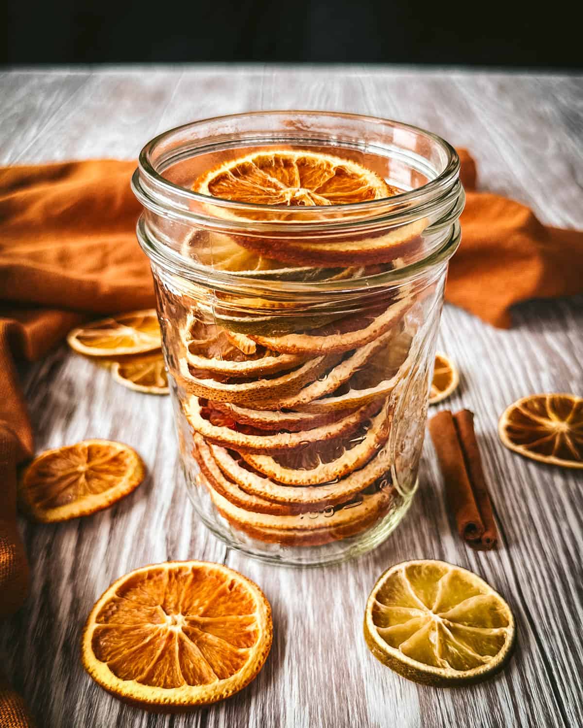 A jar of dried oranges sitting on a light gray surface, surrounded by citrus slices and an orange cloth. 