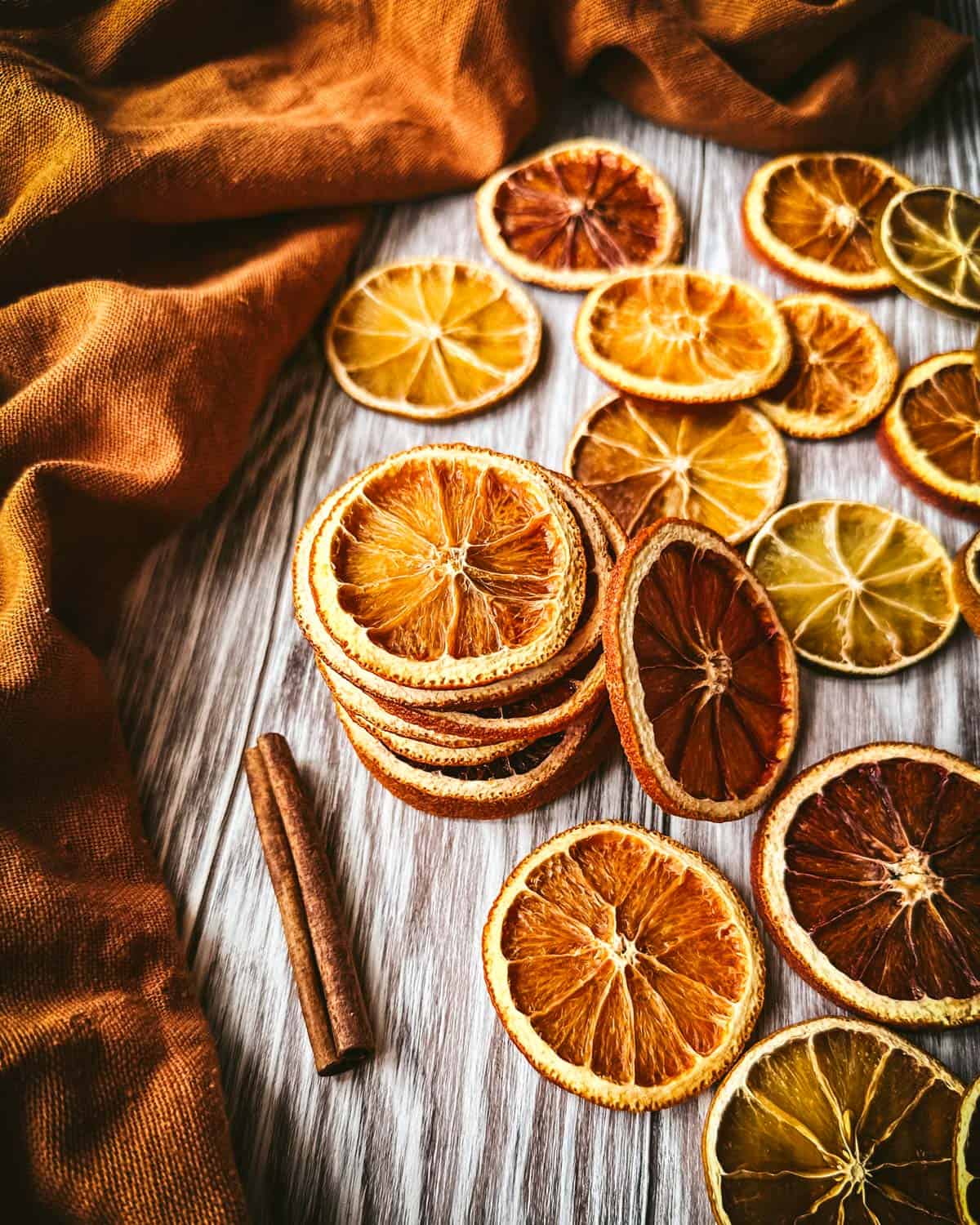 A stack of dried orange slices surrounded by orange slices and cinnamon sticks laying on a gray wood surface and a rust orange color cloth.