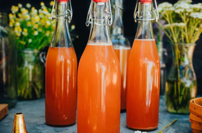 3 bottles of fermented soda surrounded by a gold funnel and fresh herbs and flowers.