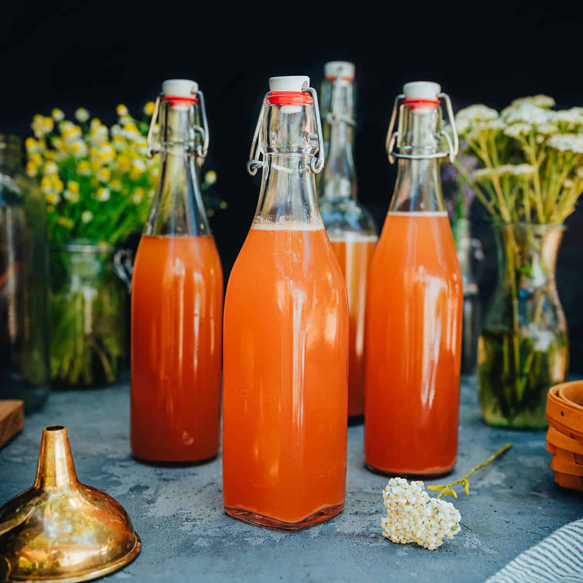 3 bottles of fermented soda surrounded by a gold funnel and fresh herbs and flowers. 