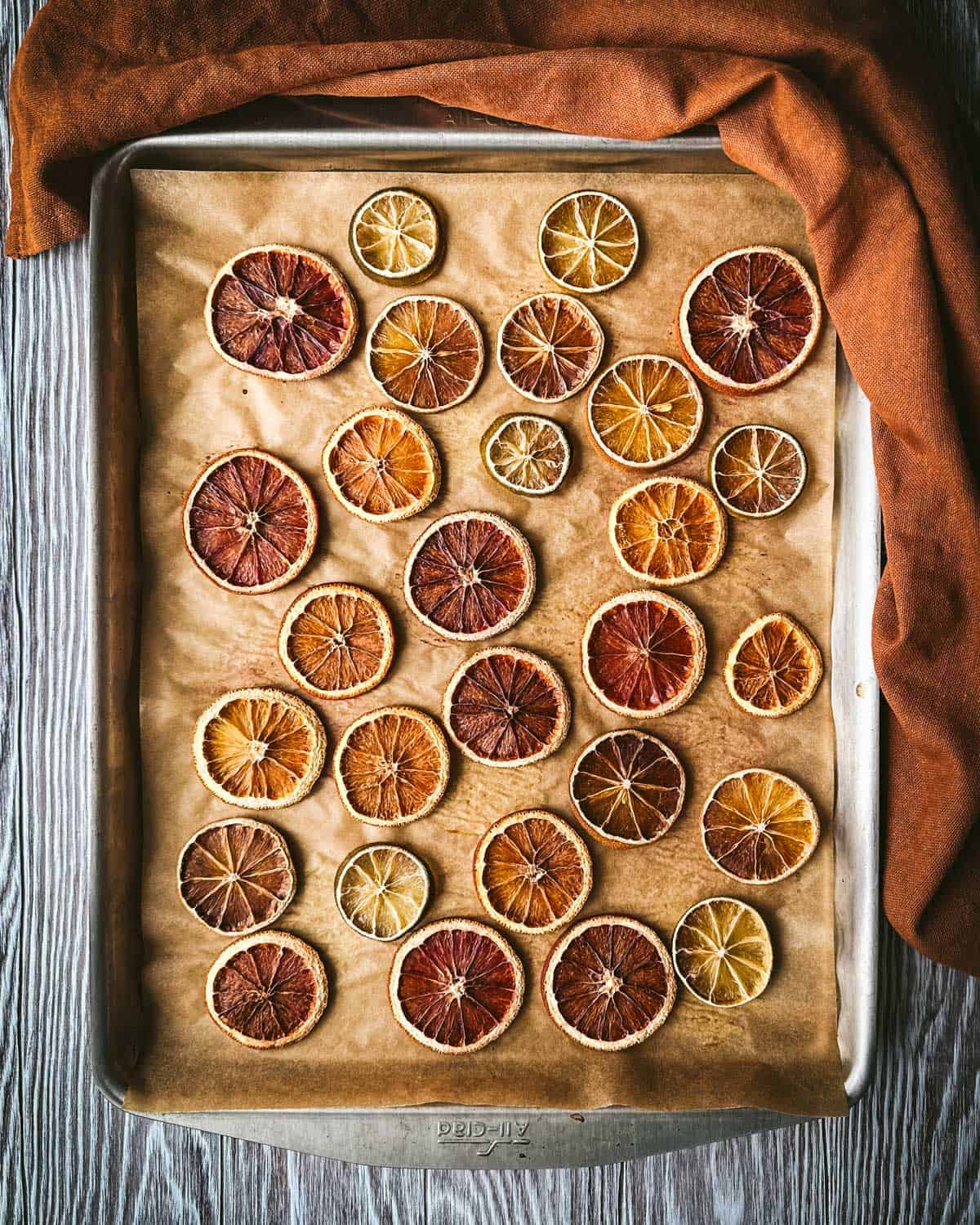 Finished dried citrus on a baking sheet, on a gray wood surface surrounded by a rust-colored cloth. 