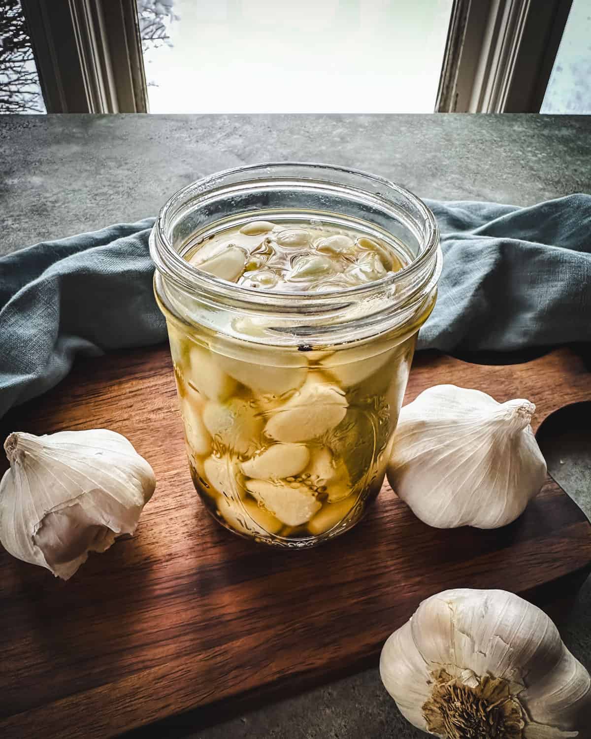 Pickled garlic in a jar on a wood cutting board, with whole garlic heads surrounding.