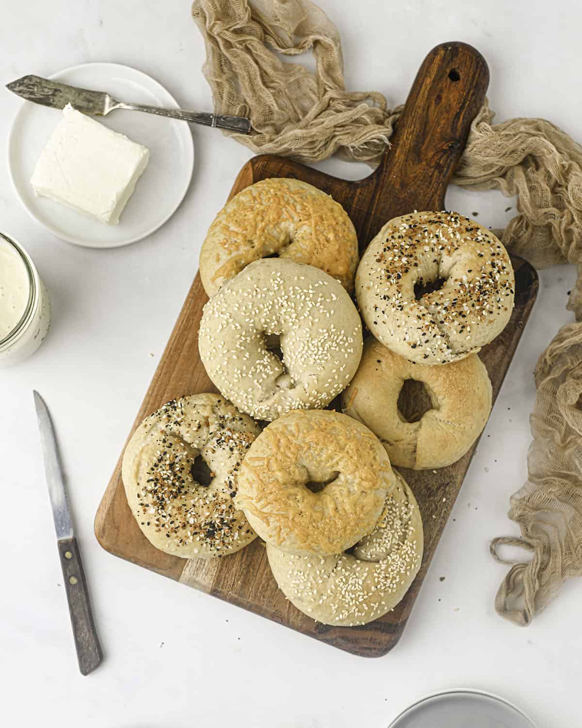 Sourdough bagels on a wood cutting board, on a white countertop surrounded by a knife and a plate of cream cheese.