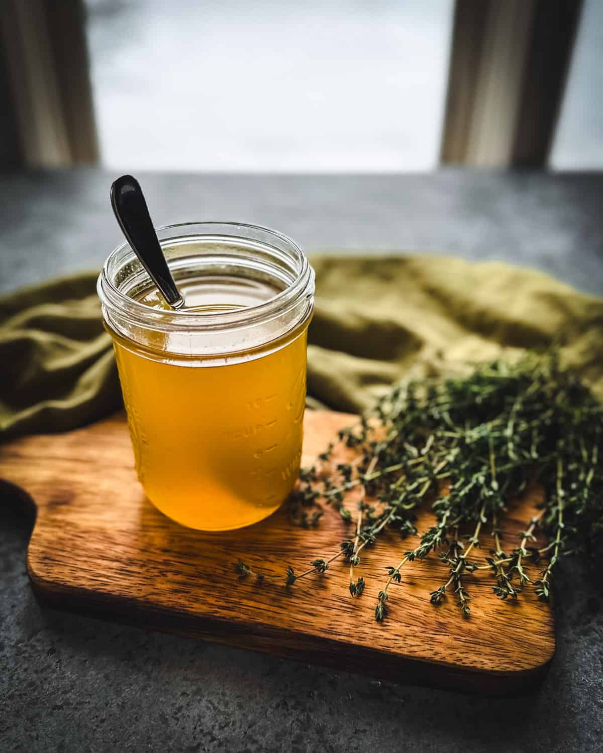 A jar of thyme cough syrup with a spoon in it, on a wood cutting board, surrounded by fresh thyme and an olive green cloth.