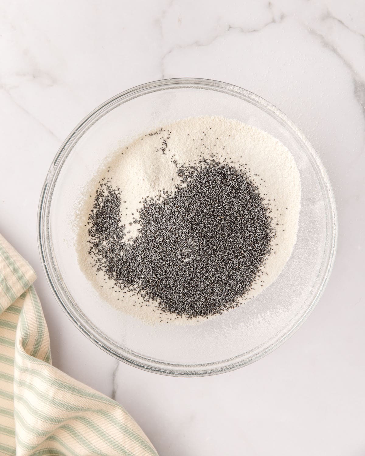 Dry ingredients and poppy seeds in a clear bowl, on a white counter with a light striped towel surrounding, top view. 