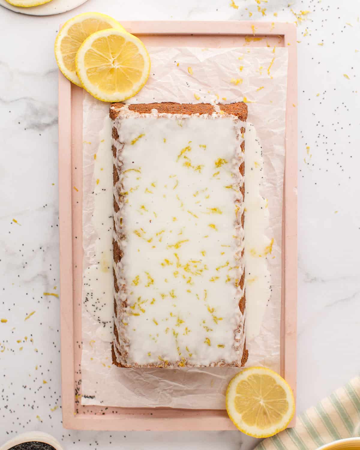 A lemon poppy seed cake with lemon glaze on it, garnished with lemon zest. Surrounded by fresh lemon slices, top view. 