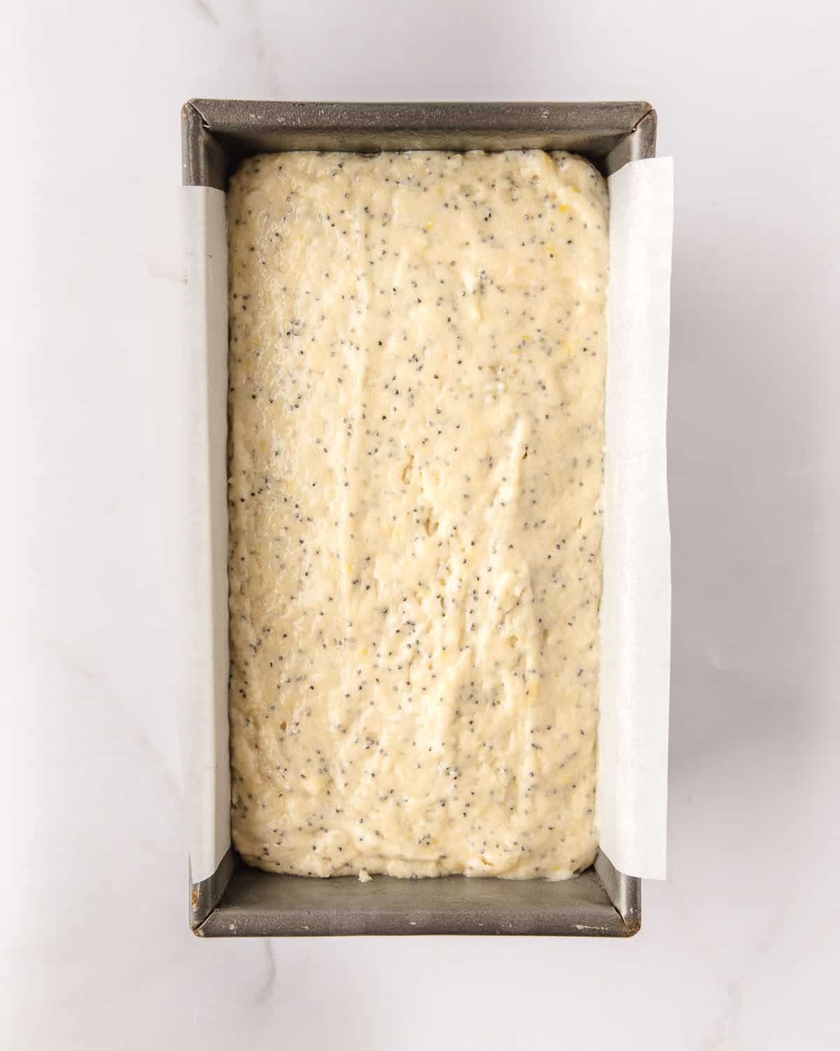 A loaf pan lined with white parchment paper, filled with lemon poppy seed loaf batter, on a white counter. 