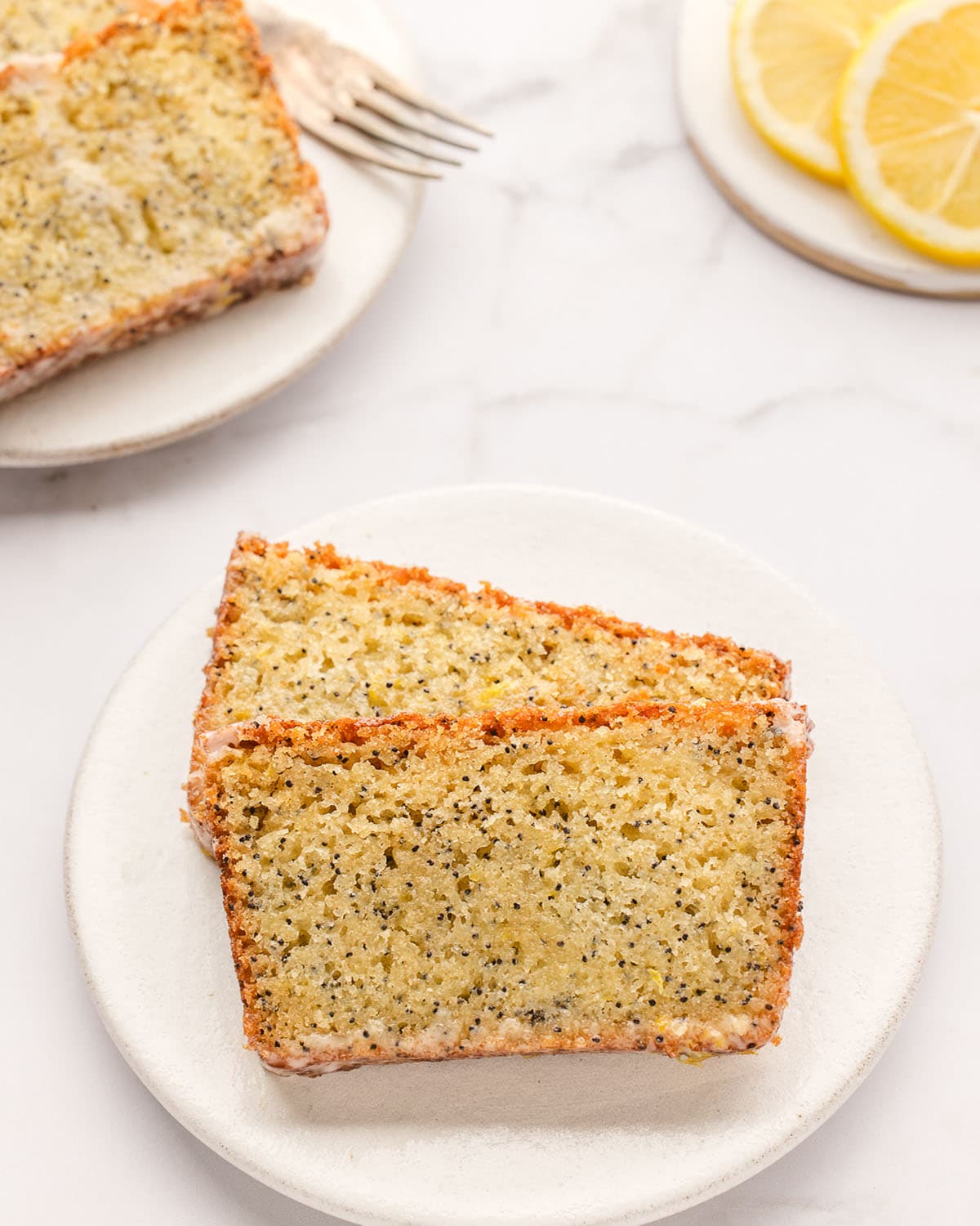 Two slices of lemon poppy seed loaf on a small white plate, on a white counter top. Surrounded by another plate with slices, and a plate of lemons. 