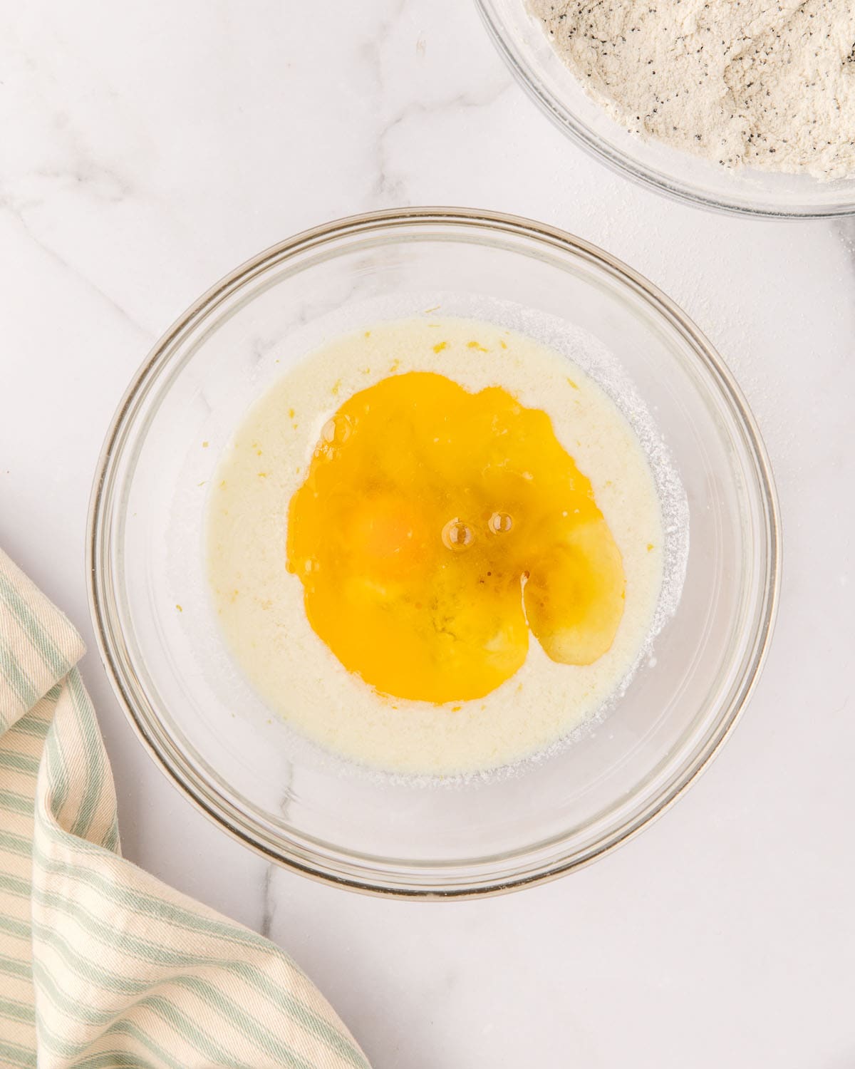 Wet ingredients in a clear bowl on a white counter, surrounded by a light striped towel, top view. 