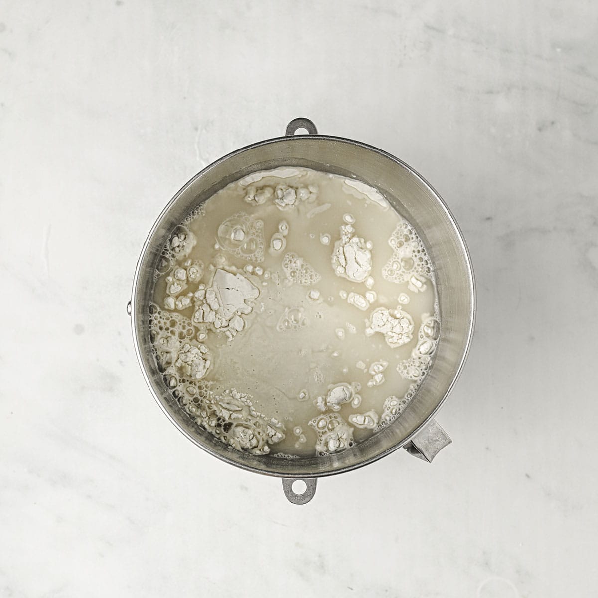 Sourdough bagel ingredients in a stand mixer bowl sitting on a white counter, top view. 