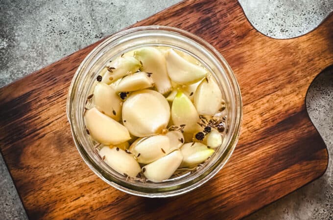 A jar filled with pickled garlic cloves, on a wood cutting board with a gray background, top view.