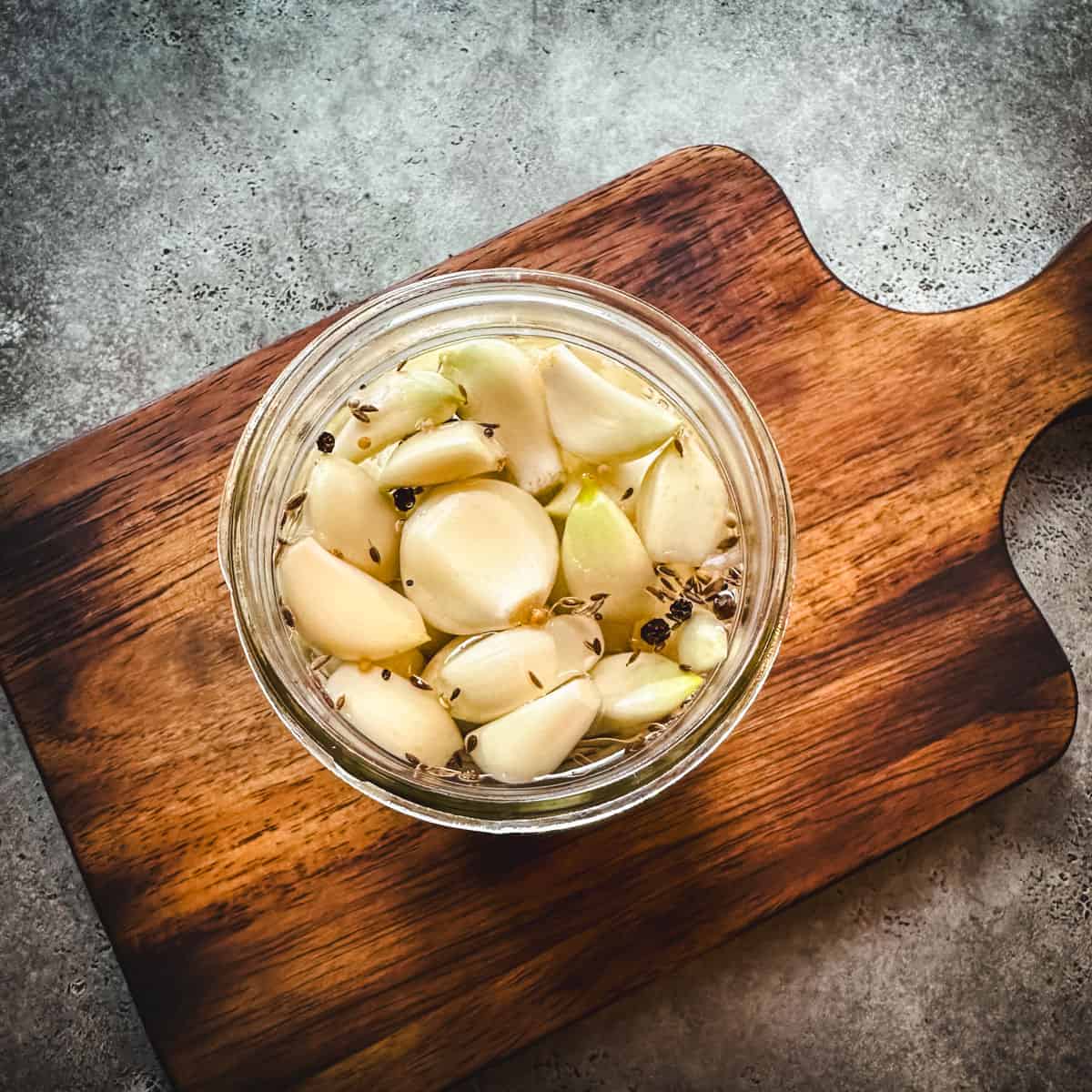 A jar filled with pickled garlic cloves, on a wood cutting board with a gray background, top view. 
