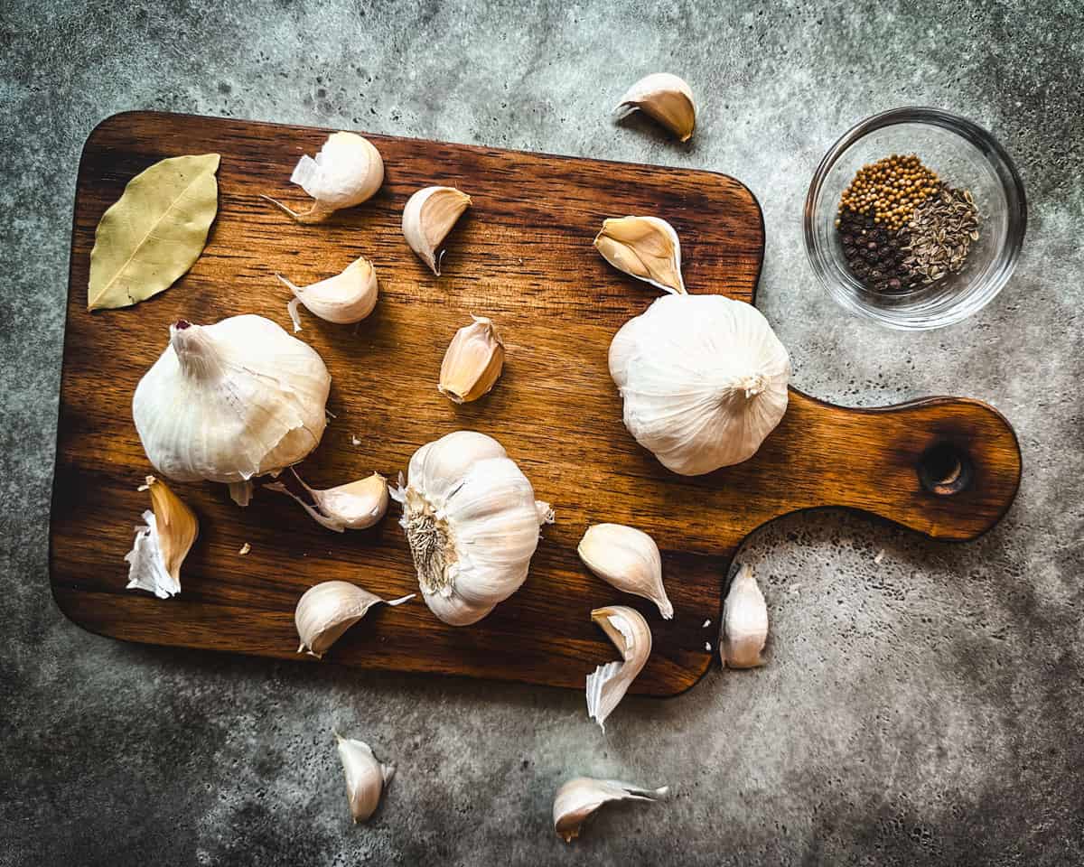 A wood cutting board with heads of garlic, cloves of garlic, a bay leaf, and a jar of pickling spices, top view. 