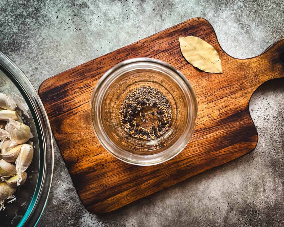 A pint sized jar with pickling spices in it, on a wood cutting board, top view. 