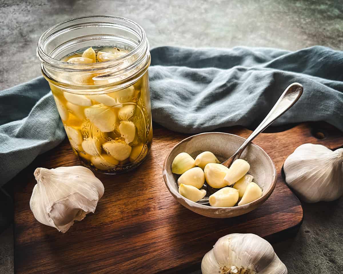 A jar and a bowl of pickled garlic with a spoon, surrounded by a blue cloth and several garlic heads. 