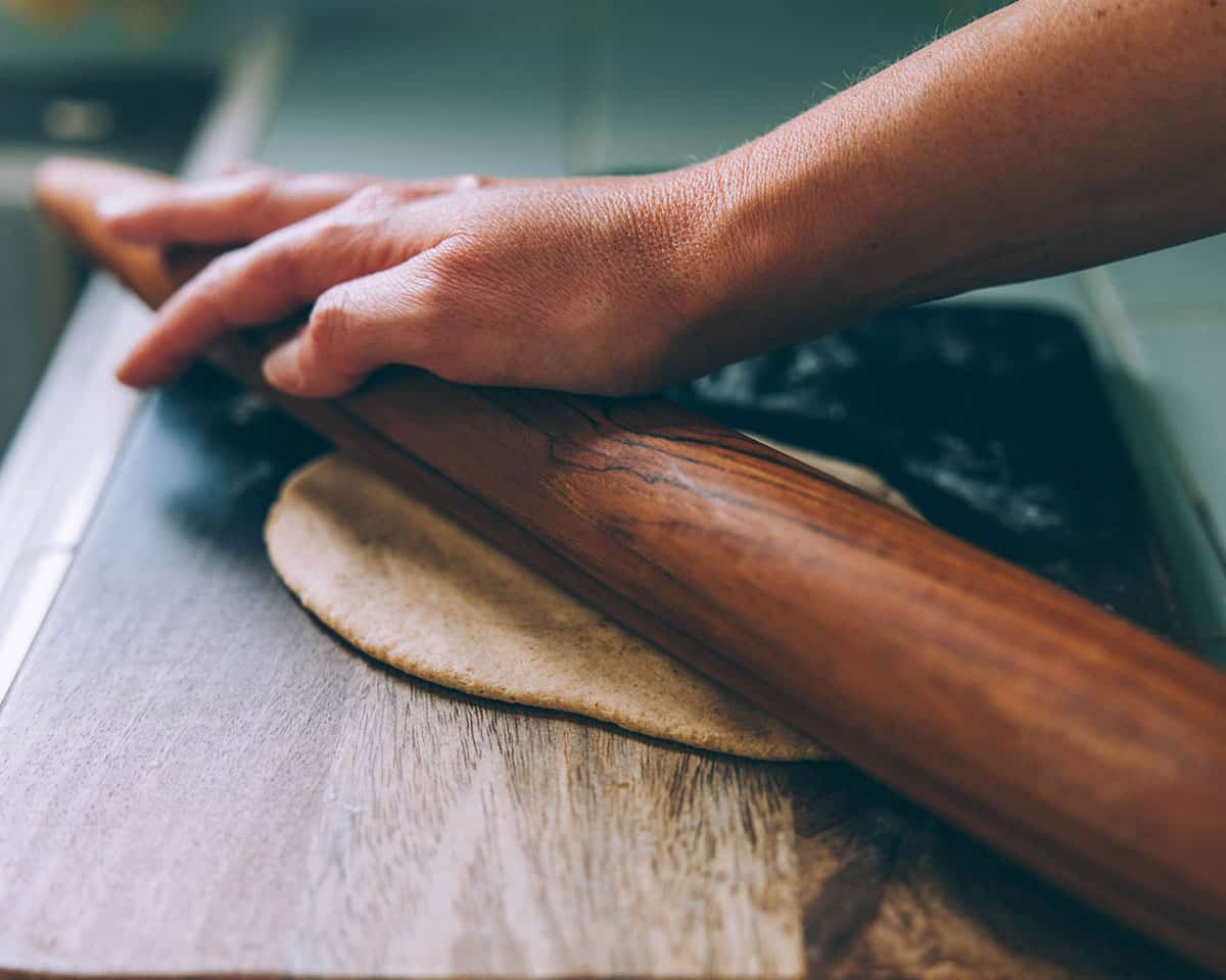 A hand using a wooden rolling pin to roll out a piece of flatbread dough onto a dark wood cutting board.