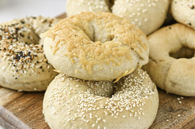 Sourdough bagels topped with sesame seeds, cheese, and everything bagel seasoning on a wood cutting board with a white counter background.