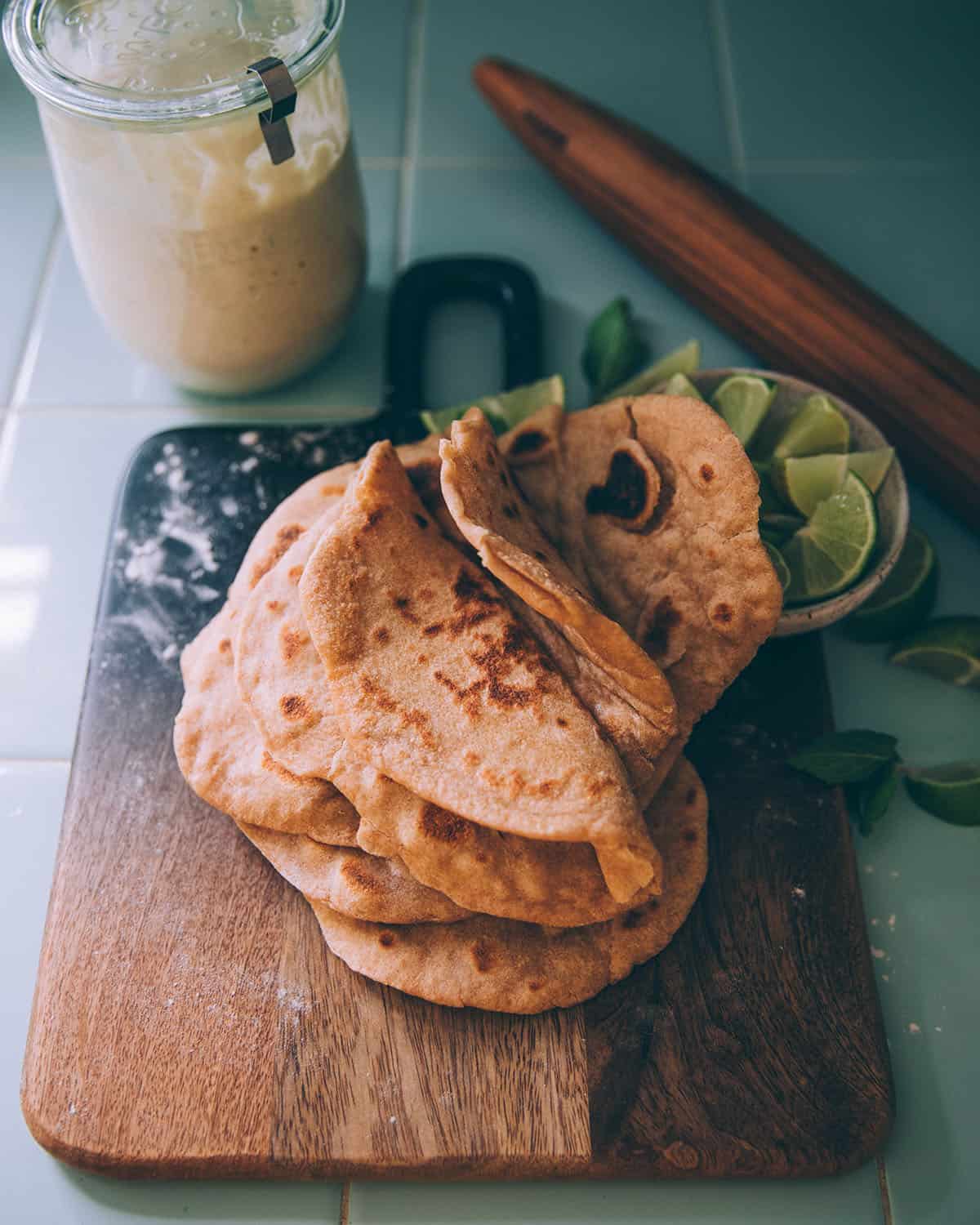 A stack of folded sourdough flatbreads on a wood cutting board, surrounded by a jar of sourdough starter and a wood rolling pin. 