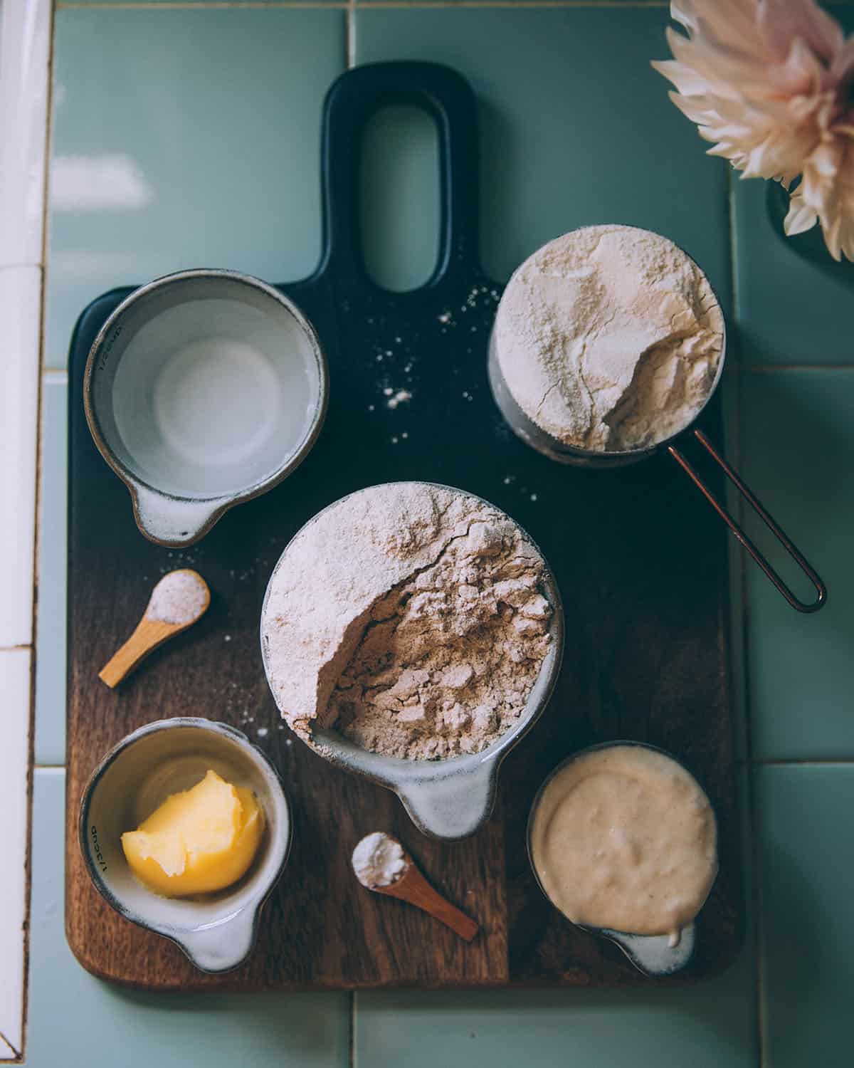 Ingredients for sourdough flatbread in bowls and measuring cups on a wood cutting board, top view. 