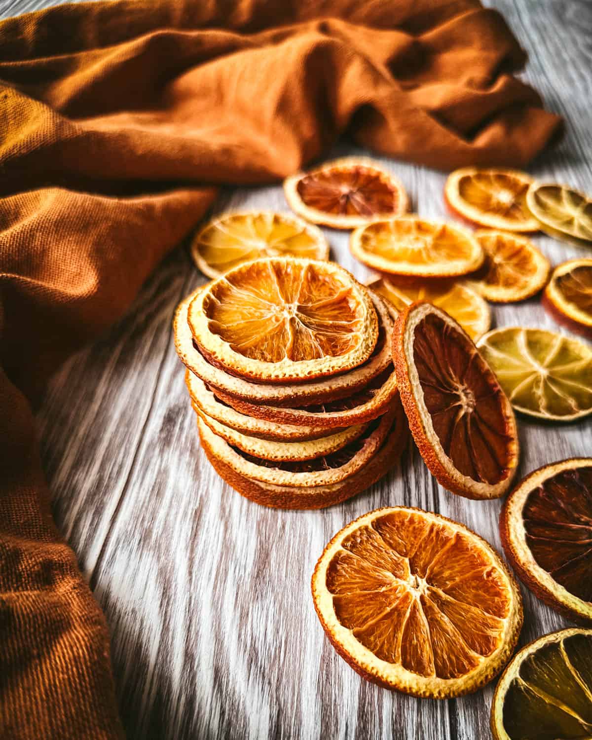 A stack of dried orange slices surrounded by orange slices laying on a gray wood surface and a rust orange color cloth. 