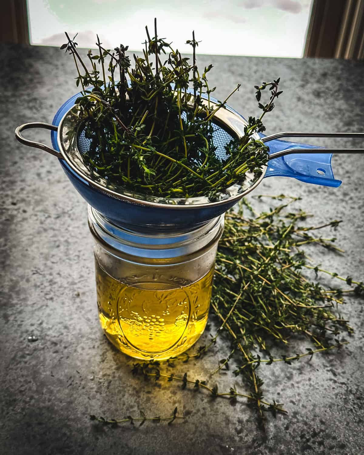 A strainer with thyme in it over a jar with the reserved thyme infusion. 