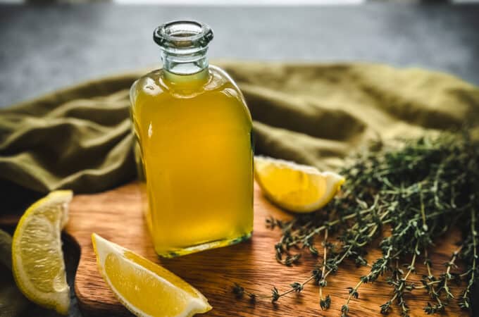 A bottle of golden colored thyme cough syrup on a wood cutting board, surrounded by lemon wedges, fresh thyme sprigs, and an olive green cloth.