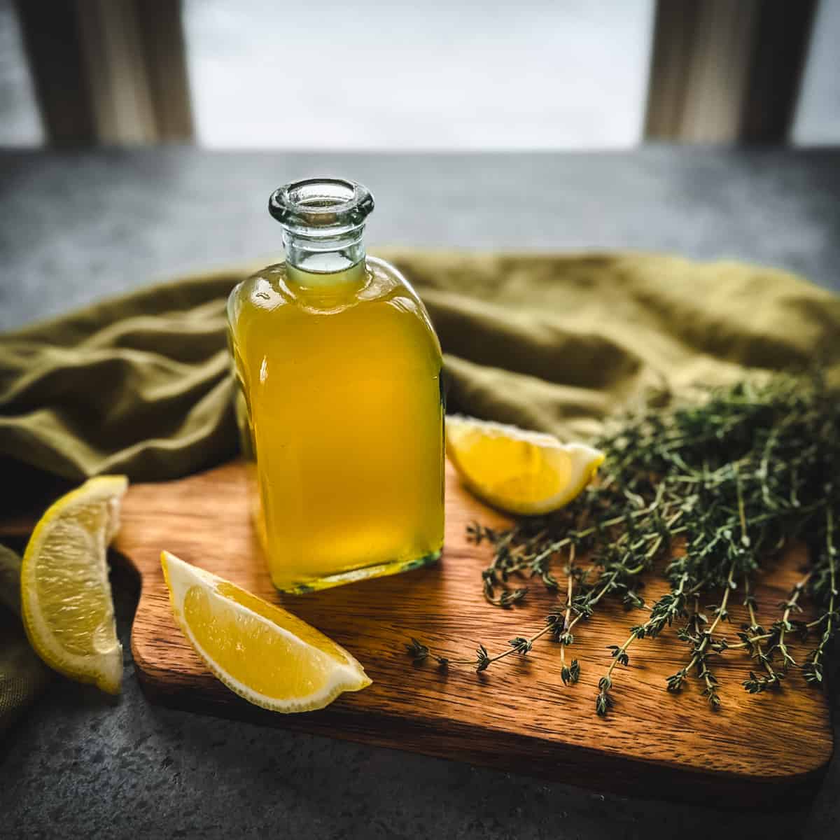 A bottle of golden colored thyme cough syrup on a wood cutting board, surrounded by lemon wedges, fresh thyme sprigs, and an olive green cloth. 