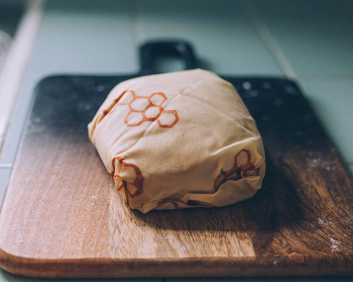 A dark wood cutting board with a dough ball wrapped in a beeswax wrap in natural light. 