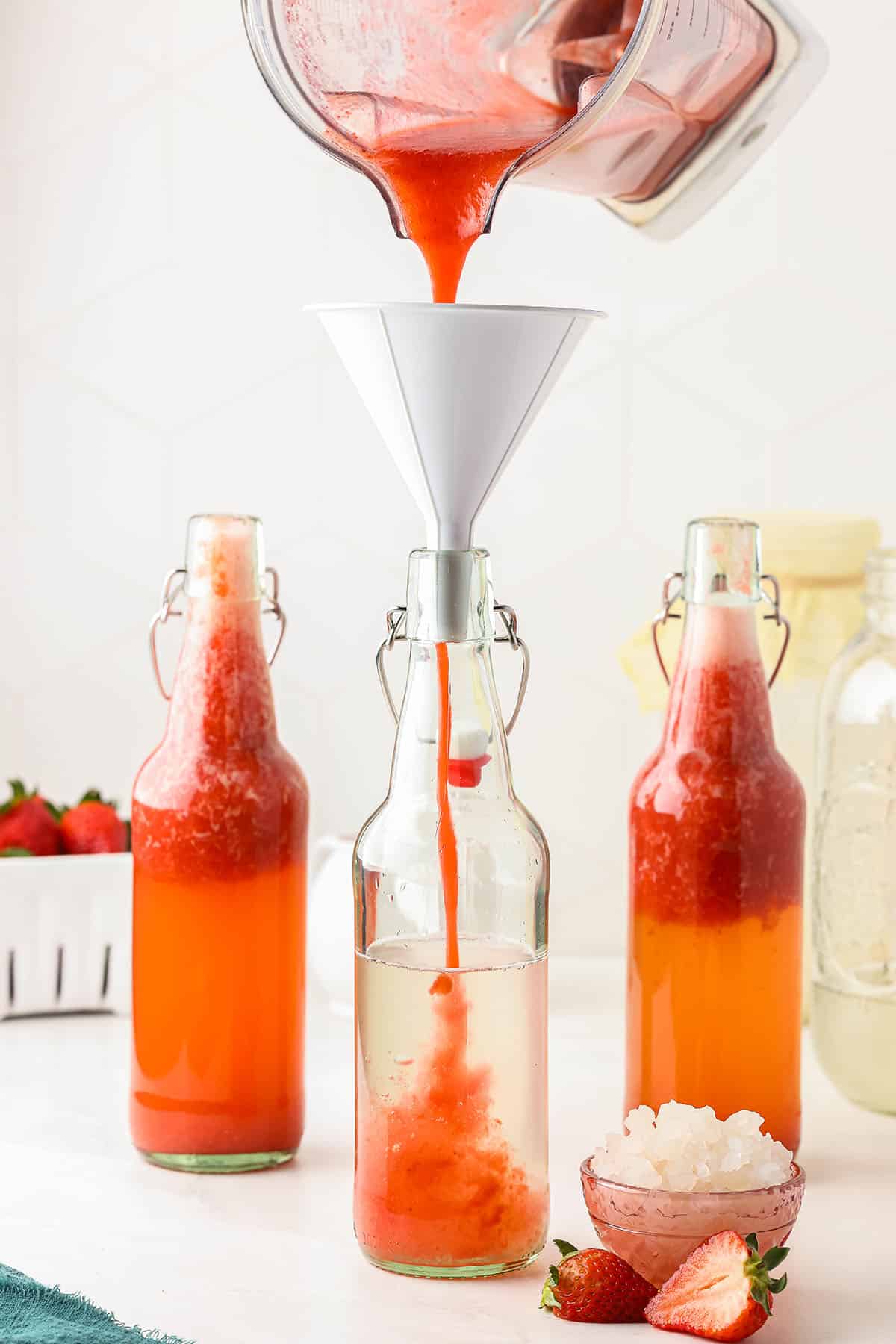 Flip top bottles filled with strawberry puree and water kefir, one with strawberry puree pouring in through a funnel, surrounded by fresh strawberries. 