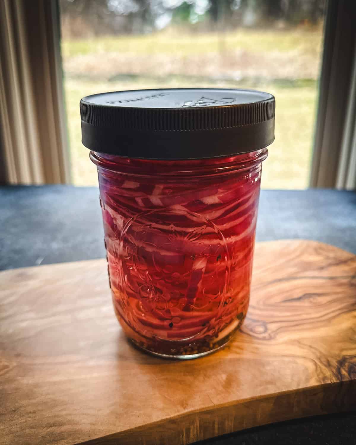 Red onions in a jar with a lid on a wood cutting board with a window in the background and natural light. 
