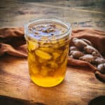 A jar of fermented ginger honey on a wood cutting board with a rust color cloth surrounding and ginger root on it.