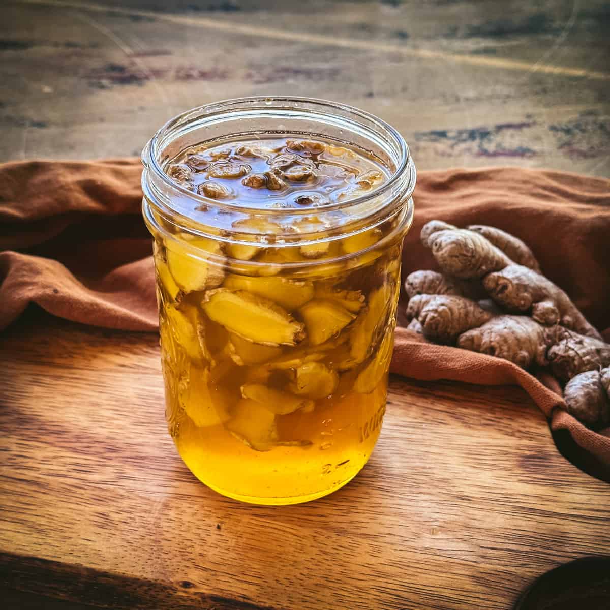 A jar of fermented ginger honey on a wood cutting board with a rust color cloth surrounding and ginger root on it. 