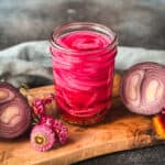A jar of beautiful pink pickled onions on a wood cutting board surrounded by a red onion cut in half and dried pink flowers.