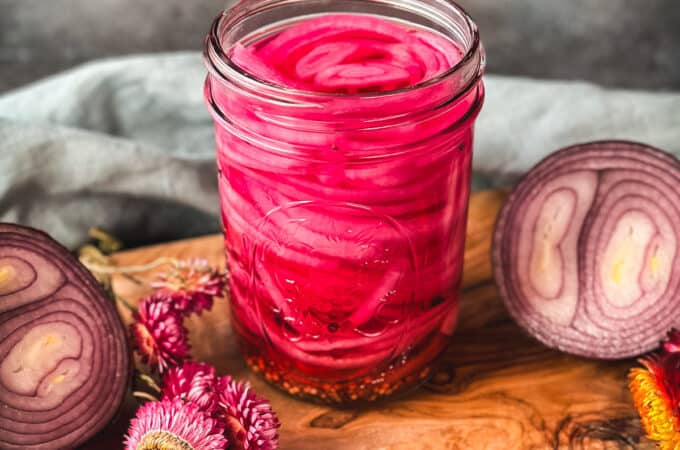 A jar of beautiful pink pickled onions on a wood cutting board surrounded by a red onion cut in half and dried pink flowers.