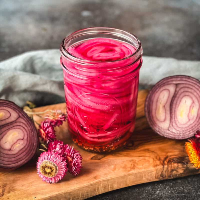 A jar of beautiful pink pickled onions on a wood cutting board surrounded by a red onion cut in half and dried pink flowers.