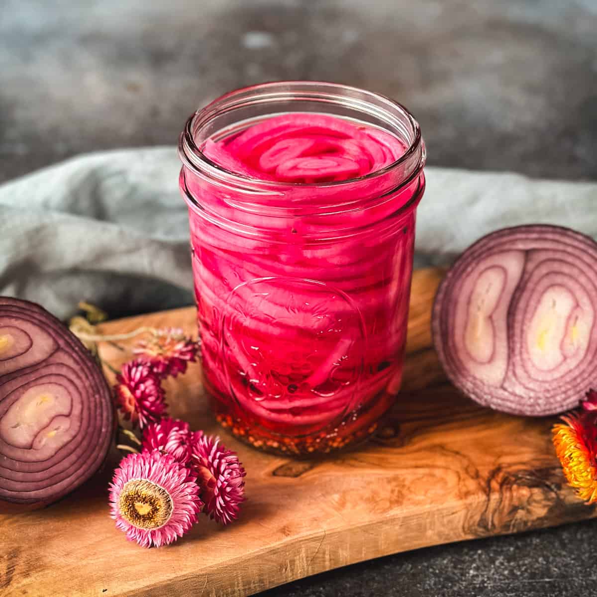 A jar of beautiful pink pickled onions on a wood cutting board surrounded by a red onion cut in half and dried pink flowers. 