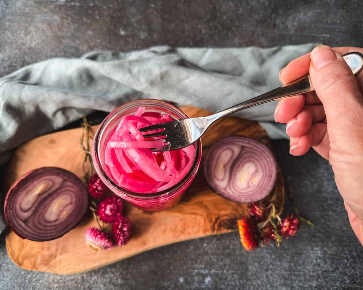 A jar of pink pickled onions with a fork lifting some up from the jar, on a wood cutting board surrounded by cut onions and dried flowers with a light blue cloth. Top view.