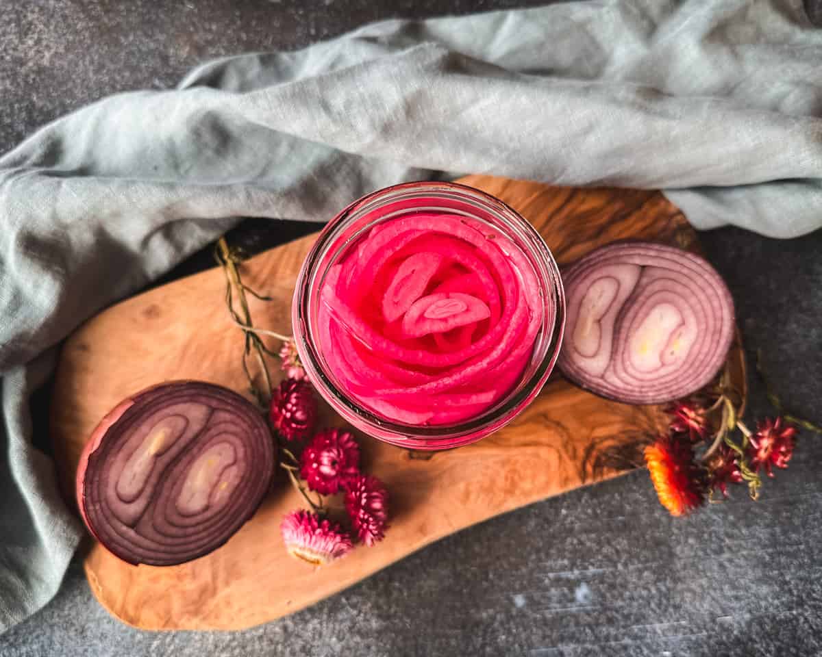 Pickled red onions in a jar on a wood cutting board surrounded by onion halves and dried flowers. Top view.