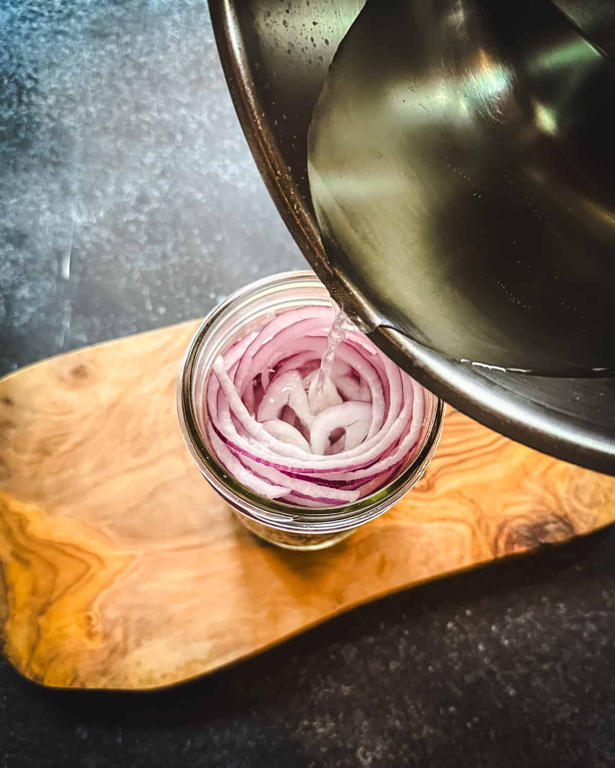 Vinegar brine pouring over the jar of onions and pickling spices, on a wood cutting board. 