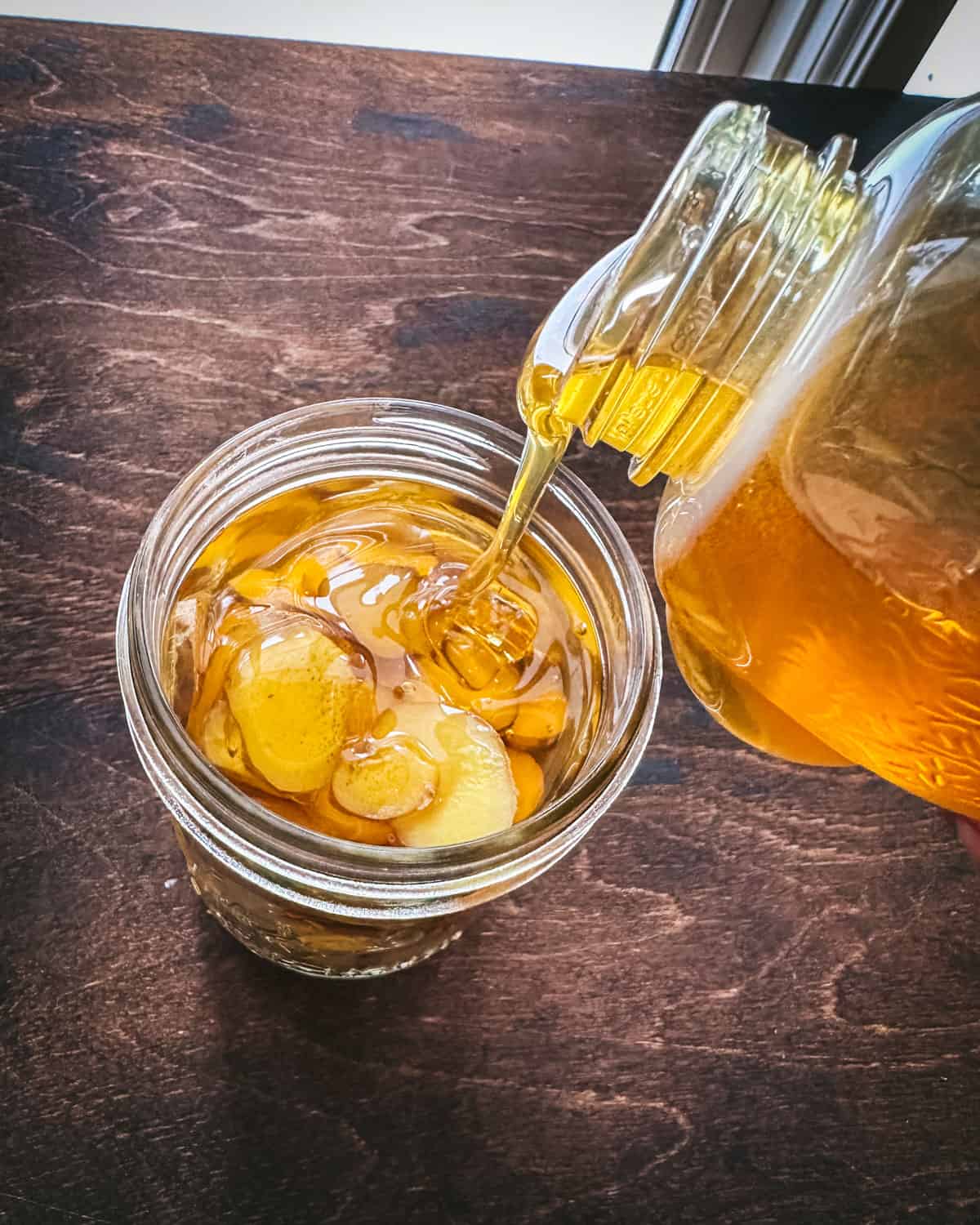 Honey pouring from a honey bear into an open jar of sliced ginger on dark wood surface, top view.