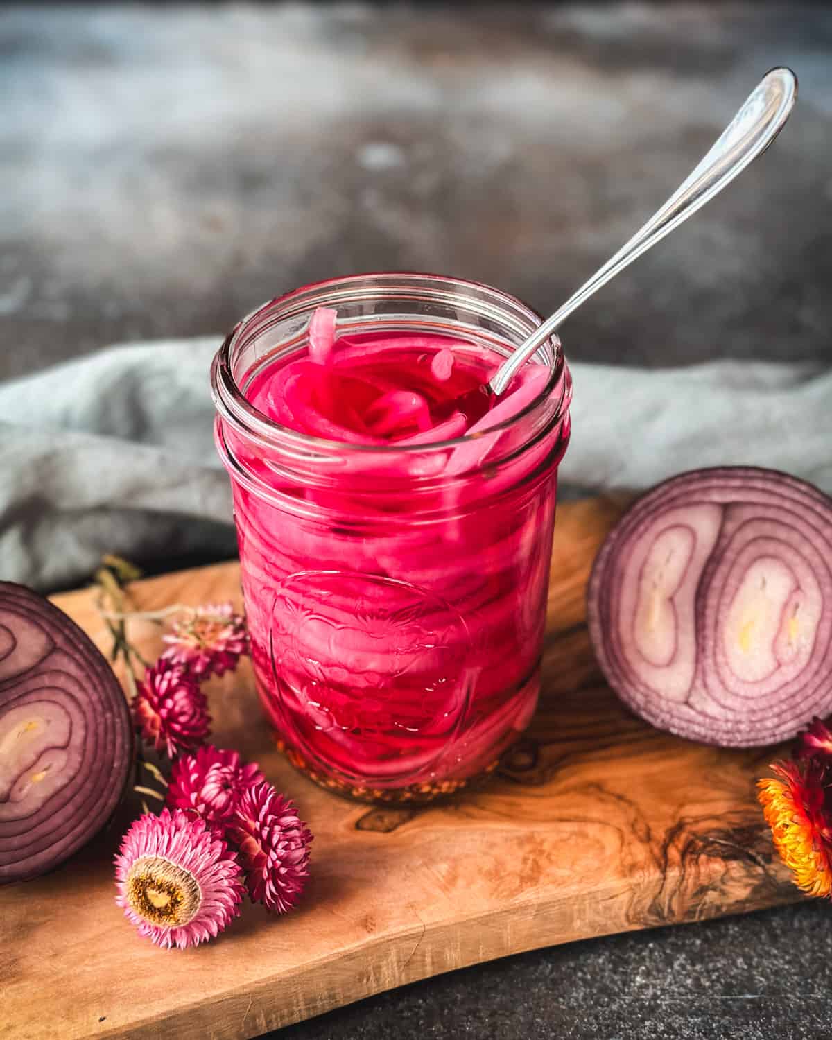 A jar of pickled red onions that are a beautiful bright pink color with a fork in it, on a wood cutting board surrounded by a cut onion and dried flowers.