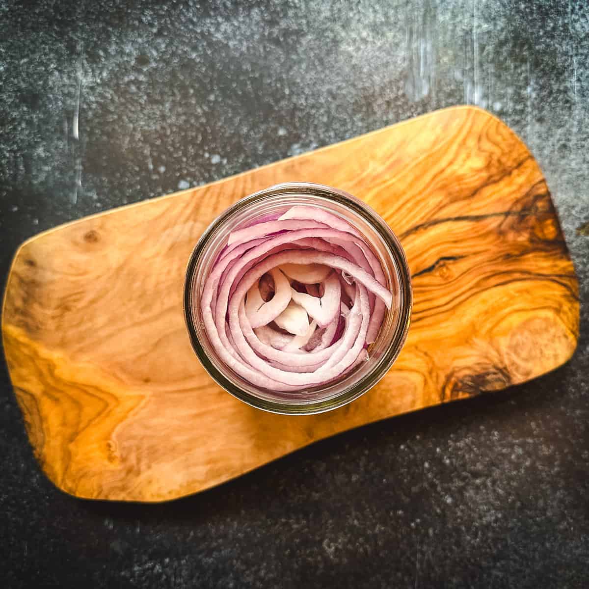 A wood cutting board with a jar of sliced onions in it, top view. 