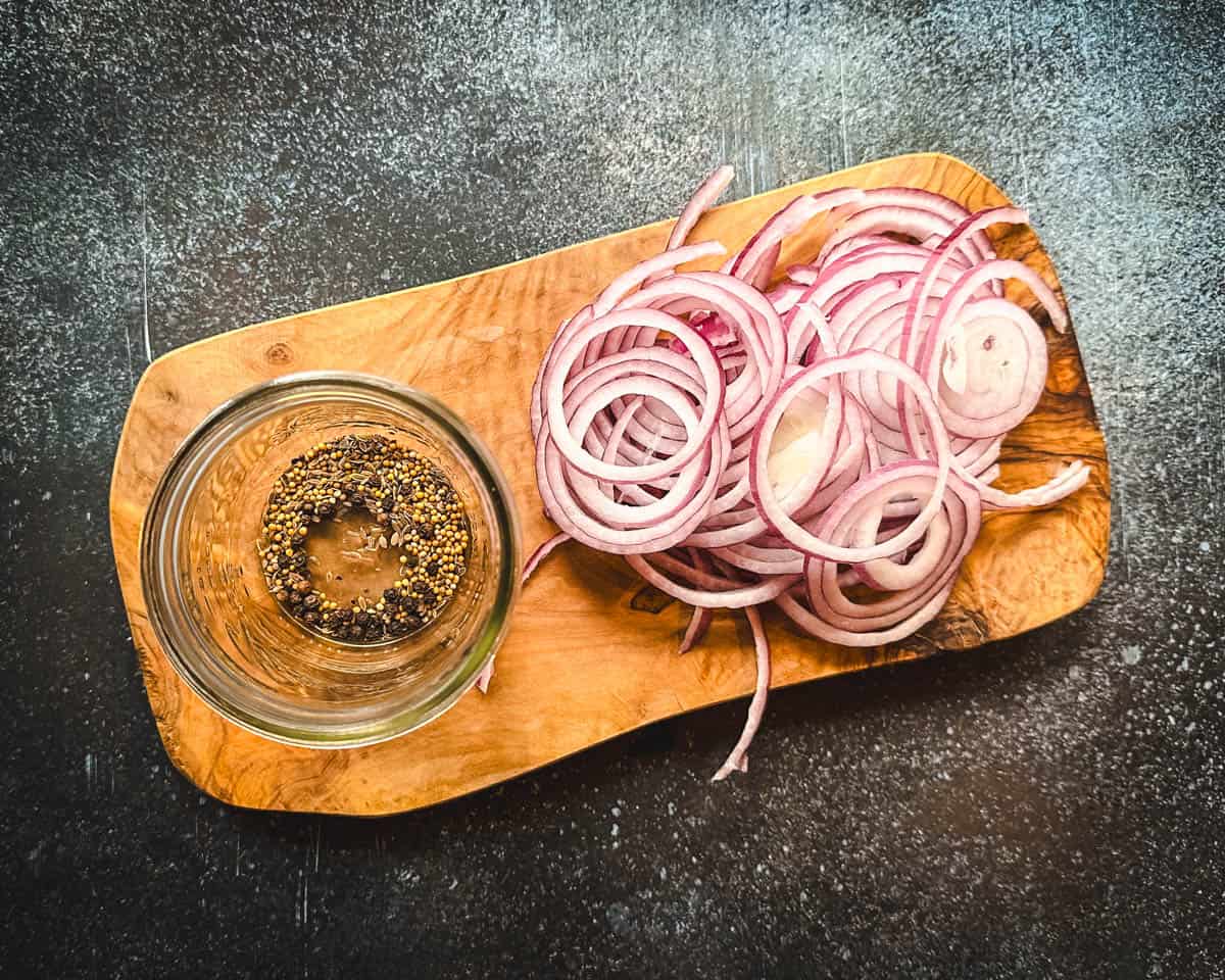 A wood cutting board with sliced red onions and a jar with pickling spices. 
