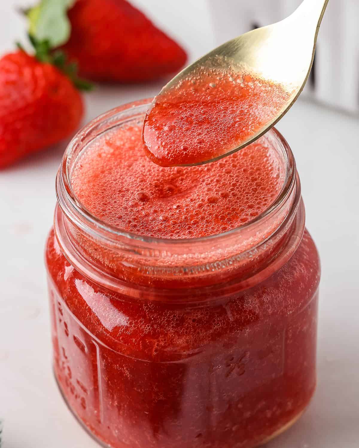 A small jar of strawberry puree with a spoon lifting some out, surrounded by fresh strawberries. 