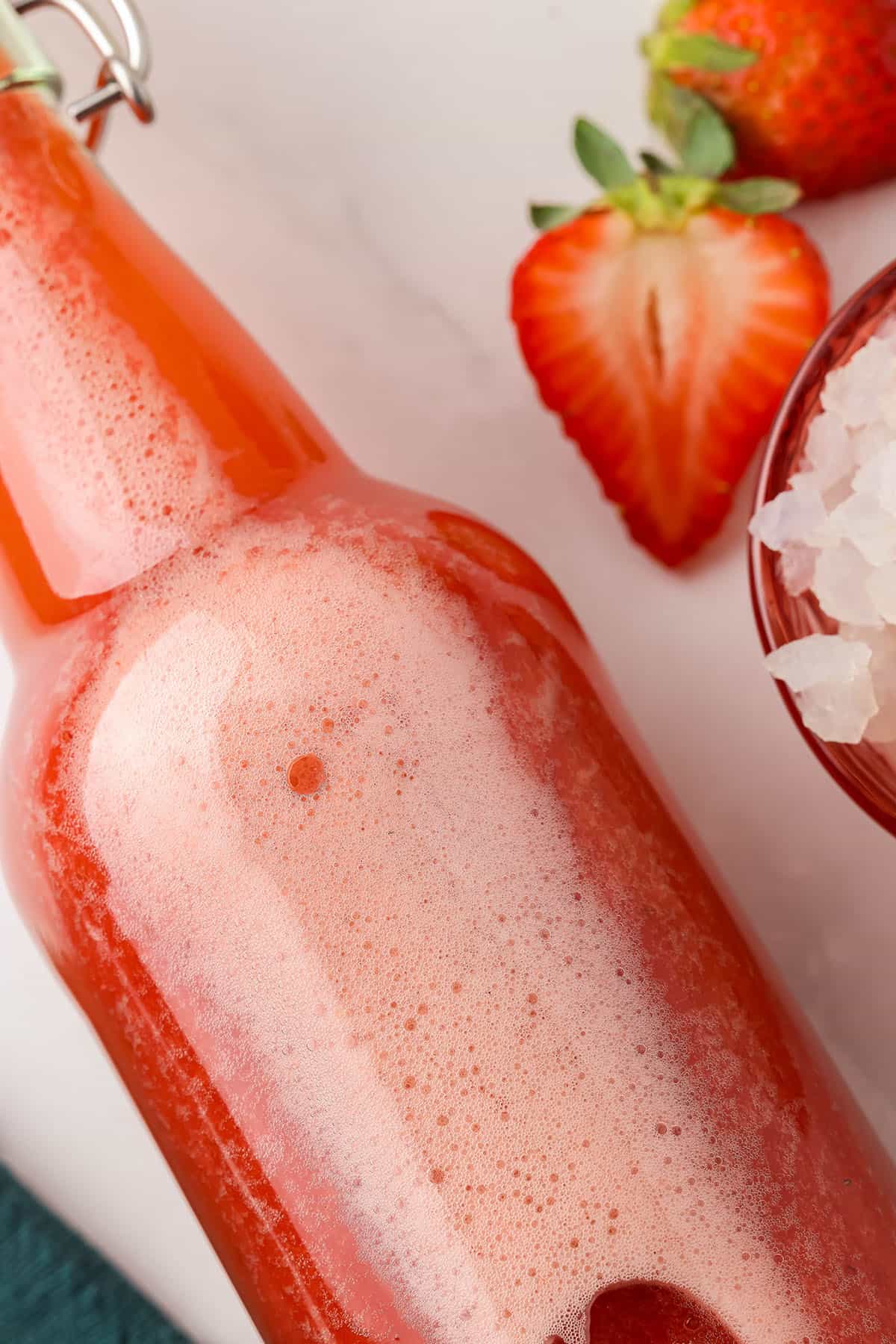 A bottle of strawberry water kefir on its side with fizz formed, surrounded by fresh strawberries. 
