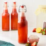 A bottle of strawberry water kefir with a flip top lid, surrounded by a small bowl of water kefir grains, fresh strawberries, lemons, and more bottles of strawberry kefir.