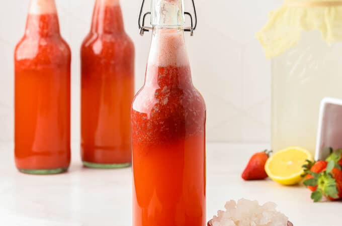 A bottle of strawberry water kefir with a flip top lid, surrounded by a small bowl of water kefir grains, fresh strawberries, lemons, and more bottles of strawberry kefir.